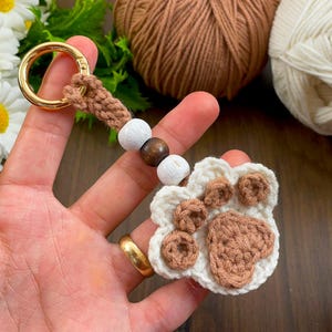Puede incluir: Un llavero de crochet hecho a mano con un diseño de huella de pata. El llavero tiene un cierre dorado, una correa de crochet marrón y una cadena de cuentas de madera blancas y marrones. La huella de pata está hecha de hilo blanco y marrón.