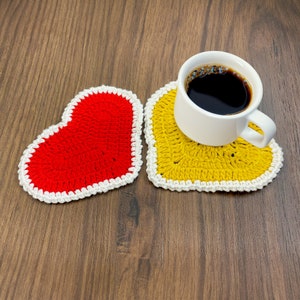 May include: Two crocheted heart-shaped coasters, one red and one yellow, with white edging. A white coffee cup with black coffee sits on the yellow coaster.