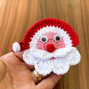 Peut inclure: Une décoration de Noël en crochet représentant un Père Noël avec un chapeau rouge, une barbe blanche et un visage rose. L'ornement a des yeux globuleux et un nez rouge.