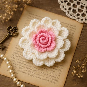 May include: A crocheted flower in cream and pink, resembling a dahlia, resting on aged paper with text. A pearl necklace and a decorative key are also present, creating a vintage aesthetic.