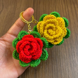 Może przedstawiać: Dwa breloki do kluczy z szydełkowymi różami. Jeden jest czerwony, drugi żółty. Oba mają zielone liście i złote kółka na klucze. Róże wykonane są z teksturowanej przędzy, a liście mają falisty brzeg.