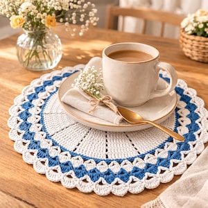 May include: A handmade, round crochet placemat in white and blue, with a scalloped edge. A beige ceramic mug filled with coffee sits on a matching saucer, with a gold spoon and a folded linen napkin. A small bouquet of flowers is tied to the napkin.