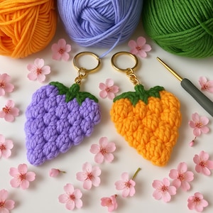 May include: Two crocheted strawberry keychains, one purple and one orange, with gold key rings. The keychains are surrounded by pink blossoms.