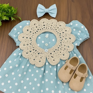 Puede incluir: Un conjunto de beb&eacute; con un vestido azul claro con lunares blancos, un cuello de ganchillo beige, un lazo a juego y zapatos Mary Jane beige. El vestido tiene mangas cortas y botones. El cuello de ganchillo tiene forma de flor.