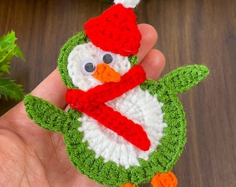 Könnte beinhalten: Ein gehäkeltes Pinguin-Ornament mit roter Mütze und Schal. Der Pinguin ist grün und weiß mit orangenen Füßen.