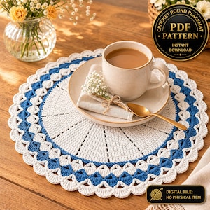 May include: A round crochet placemat featuring a white center with radiating stitches and a decorative border of alternating blue and white scalloped loops. A ceramic mug, a folded napkin, and a gold spoon rest on the placemat. Text reads PDF PATTERN.