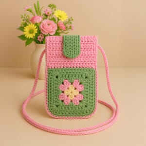 Puede incluir: Una funda de teléfono de crochet rosa con un diseño de flor de crochet verde y una correa rosa.