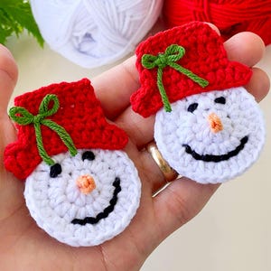 Puede incluir: Dos adornos de muñeco de nieve de crochet con sombreros rojos y lazos verdes. Los muñecos de nieve tienen caras blancas con ojos negros, sonrisas negras y narices naranjas.