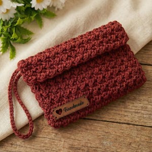May include: A handmade, rust-colored crochet clutch with a flap closure and a wrist strap. The clutch features a textured stitch pattern and a small leather tag that reads "Handmade" with a heart symbol.