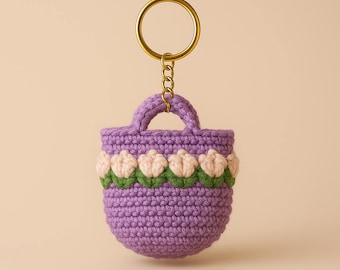 May include: A purple crocheted bag keychain with a white and green flower design. The bag has a small handle and a gold metal key ring.