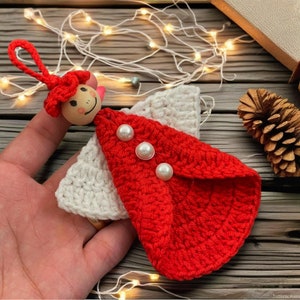 May include: A crocheted angel ornament with a red dress and white wings. The angel has a wooden head with a painted face and red yarn hair.