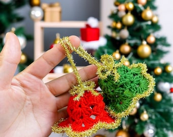 May include: Two crocheted Christmas ornaments in the shape of bells. One bell is red with gold trim and the other is green with gold trim. The ornaments are held in a hand against a blurred background of a Christmas tree.