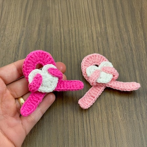 May include: Two handmade crochet heart-shaped appliques. One is held in a hand. The appliques feature a bright pink heart with a white heart in the center. The other applique is light pink with a white heart.