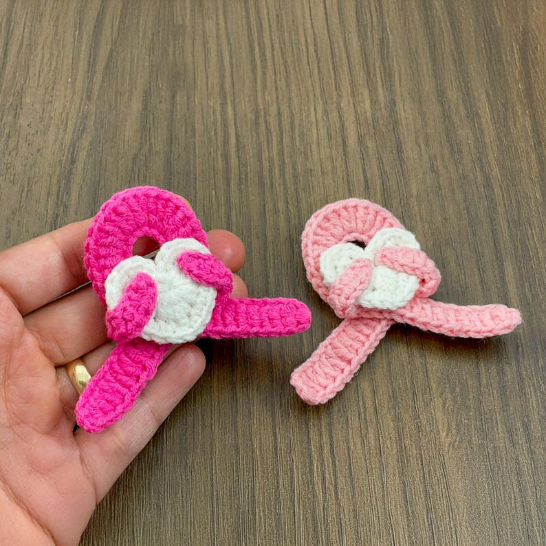 May include: Two handmade crochet heart-shaped appliques. One is held in a hand. The appliques feature a bright pink heart with a white heart in the center. The other applique is light pink with a white heart.