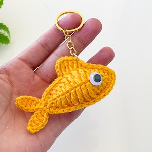 May include: A handmade, yellow crochet fish keychain with a gold-colored keyring. The fish has a single googly eye and detailed crochet work, showcasing a textured pattern. The keychain is held in a hand against a plain background.