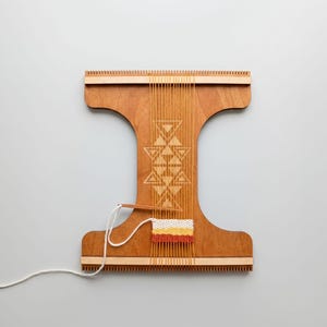 Op de afbeelding: Een houten weefgetouw met een geometrisch ontwerp in het hout geëtst. Het weefgetouw heeft verticale draden en een klein geweven gedeelte in tinten geel, oranje en wit. Een wit garen is bevestigd.