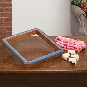 May include: A wooden embroidery frame with blue fabric edges and corner pieces. A pink, fluffy headband with yellow and red spots is next to the frame. The items are on a wooden surface.