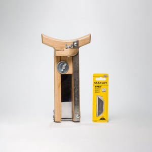 Op de afbeelding: Een houten leersplijter met een bevestigde metalen liniaal en een vleugelmoer-verstelmechanisme. Daarnaast staat een gele Stanley 1992 verpakking met vijf reservemesjes, met een hoogte van ongeveer 23 cm.