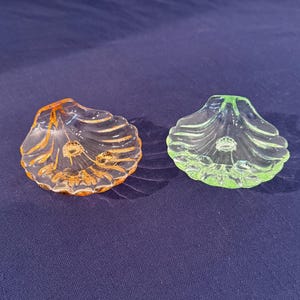 May include: Two small, decorative glass seashell dishes. One is orange, the other is green. Both have a ridged design and a small, central indentation. The dishes are likely used for holding small items or as decorative accents.
