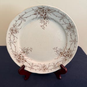 May include: A round ceramic plate with a cream-colored base and a brown transferware pattern. The design features delicate floral bouquets and thin, trailing vine garlands that encircle the rim and extend toward the center of the plate.