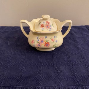 May include: A white ceramic sugar bowl with a lid, featuring a floral design in pink, blue, and red. The bowl has two handles and a scalloped rim.