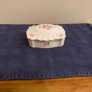 May include: A white porcelain trinket box with a scalloped lid. The lid is decorated with a floral design in shades of pink, orange, and green. The box has a gold trim and sits on a blue surface.