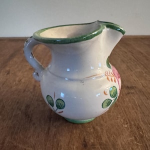 May include: A small, white ceramic pitcher with a handle and spout. The pitcher is decorated with green trim and floral designs in pink, green, and orange. The pitcher is approximately 4 inches tall.