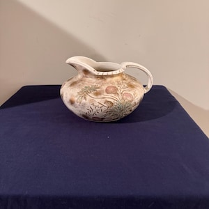 May include: A white ceramic pitcher with a brown and gold floral design. The pitcher has a wide mouth and a handle.