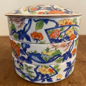 May include: A three-tiered ceramic serving dish with a white background and a floral pattern in shades of blue, red, yellow, and green. The lid has a gold trim.
