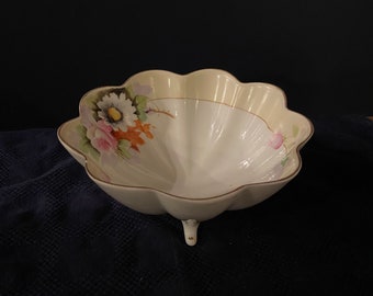 May include: A white porcelain bowl with a scalloped rim and floral design. The bowl has three legs and is decorated with pink, yellow, and orange flowers.