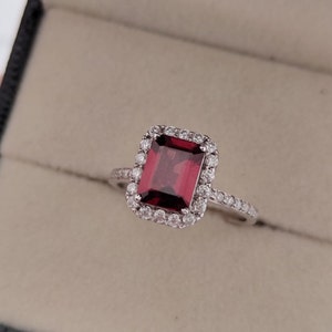 May include: A silver ring with a large, red, emerald-cut gemstone surrounded by a halo of smaller, clear gemstones.
