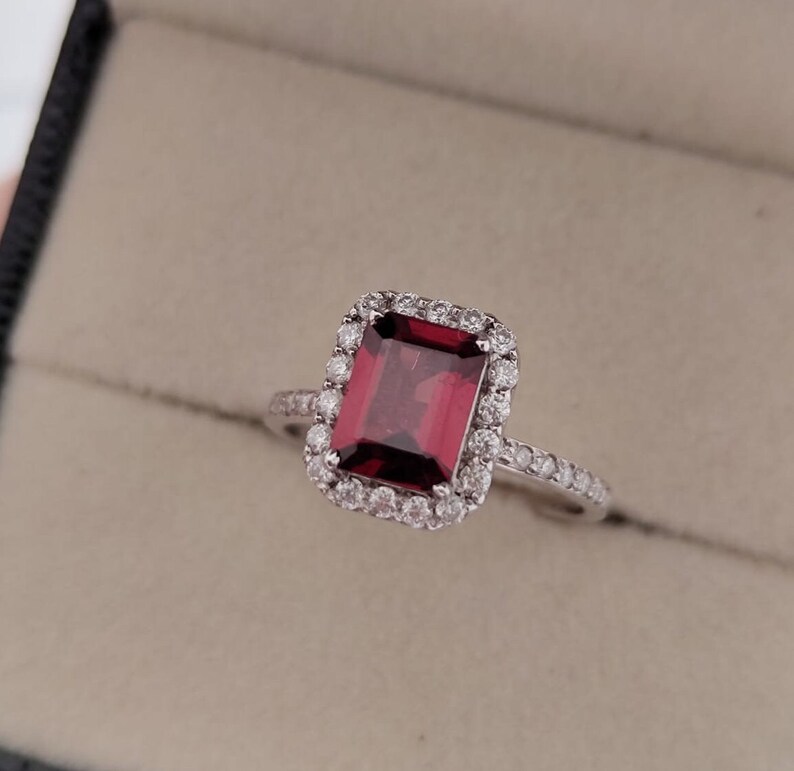 May include: A silver ring with a large, red, emerald-cut gemstone surrounded by a halo of smaller, clear gemstones.