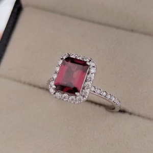May include: A silver ring with a large, square, red gemstone surrounded by a halo of smaller diamonds. The band is also set with smaller diamonds.