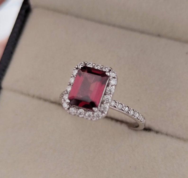 May include: A silver ring with a large, square, red gemstone surrounded by a halo of smaller diamonds. The band is also set with smaller diamonds.