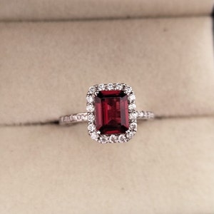 May include: A red gemstone ring with a halo of smaller diamonds. The ring is set in white metal.