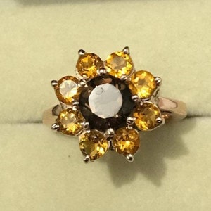 Puede incluir: Un anillo dorado con un diseño floral. La piedra central es de color marrón oscuro, rodeada de diez piedras más pequeñas de color amarillo dorado. La banda del anillo es de color oro claro.