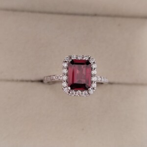 May include: A red gemstone ring with a square-shaped stone surrounded by a halo of smaller diamonds. The ring is set in white metal.