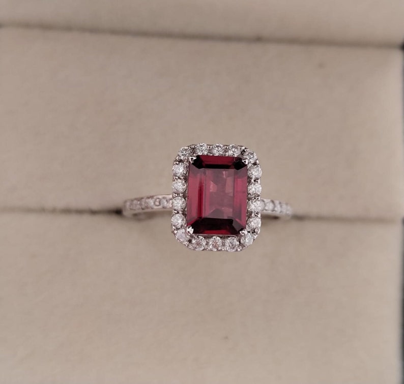 May include: A red gemstone ring with a square-shaped stone surrounded by a halo of smaller diamonds. The ring is set in white metal.