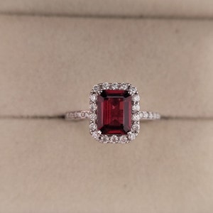 May include: A red gemstone ring with a halo of smaller diamonds. The ring is set in a silver band.