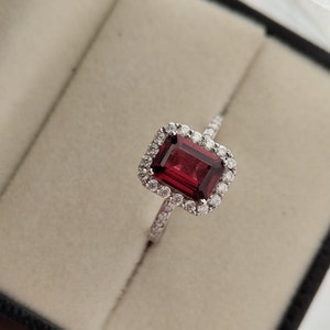 May include: A silver ring with a large, emerald-cut red gemstone surrounded by a halo of smaller diamonds.