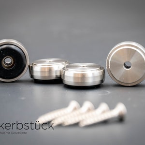 May include: Four silver metal furniture feet with black rubber pads and four silver screws. The feet have a raised lip around the edge and a small hole in the center. The screws are about 2 inches long.