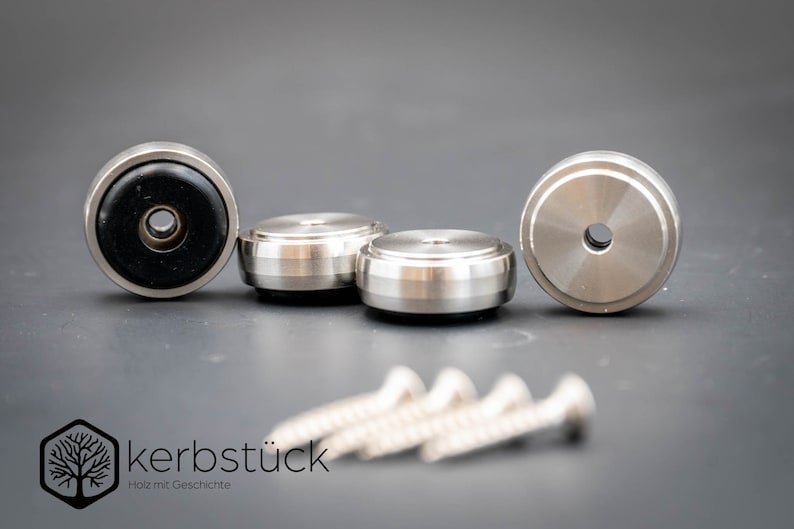 May include: Four silver metal furniture feet with black rubber pads and four silver screws. The feet have a raised lip around the edge and a small hole in the center. The screws are about 2 inches long.