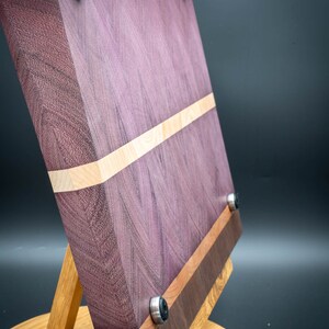 May include: A purple, walnut, and maple wood cutting board with a stand. The board has a natural wood finish and is accented with black rubber feet.