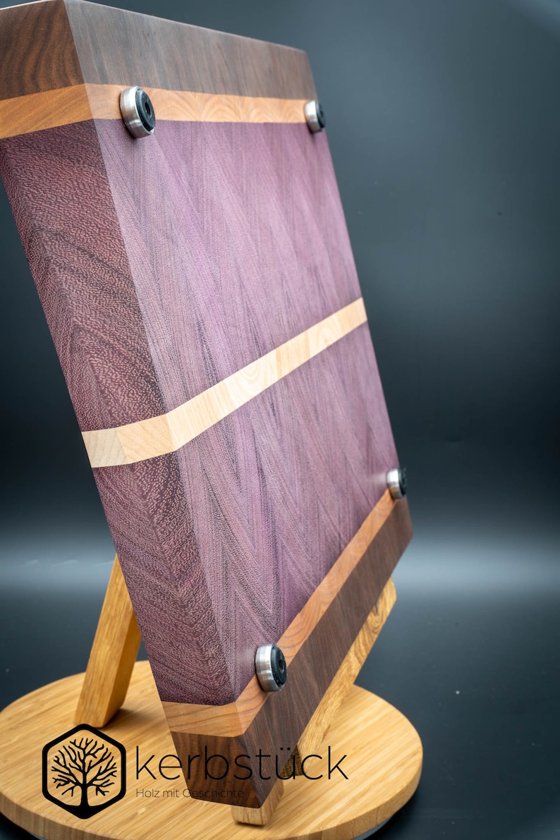 May include: A purple, walnut, and maple wood cutting board with a stand. The board has a natural wood finish and is accented with black rubber feet.