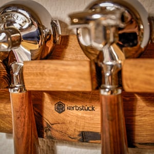 May include: A wooden espresso machine portafilter holder with two chrome portafilters. The holder is made of light brown wood and features the text "kerbstück" and a logo. The portafilters have wooden handles.