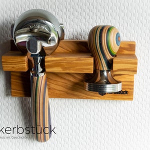 May include: Wooden espresso machine portafilter and tamper holder. The holder is made of light brown wood and holds a chrome portafilter and a tamper with a colorful striped wooden handle. The logo "kerbstück" is visible.