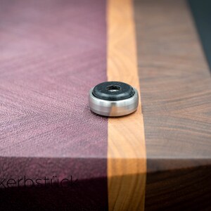 May include: A close-up of a wooden cutting board with a purple, orange, and brown wood grain pattern. The board has a metal foot with a black rubber pad on the bottom.