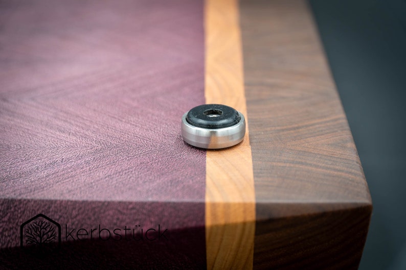 May include: A close-up of a wooden cutting board with a purple, orange, and brown wood grain pattern. The board has a metal foot with a black rubber pad on the bottom.