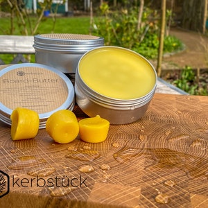 May include: Three silver tins of Board Butter, a wood care product, on a wooden cutting board. The tins are labeled "Board Butter" and "kerbstück Holz mit Geschichte". There are three yellow wax blocks on the board.