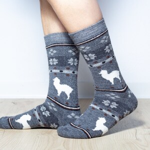 Pu&ograve; includere: Un paio di calzini grigi con un motivo di alpaca bianco. I calzini hanno una striscia marrone intorno alla parte superiore e un motivo a fiocco di neve bianco.